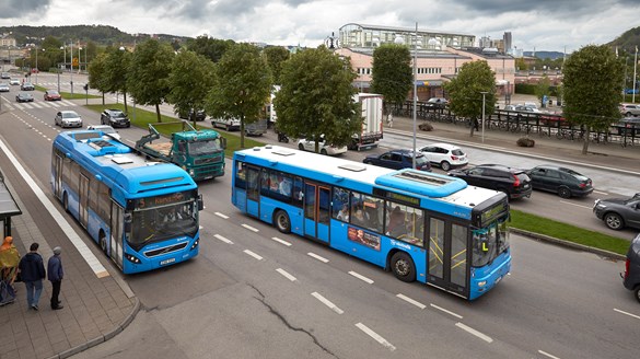Två blåa bussar på en trafikerad gata. En av dem har stannat vid en hållplats. erad gata. 